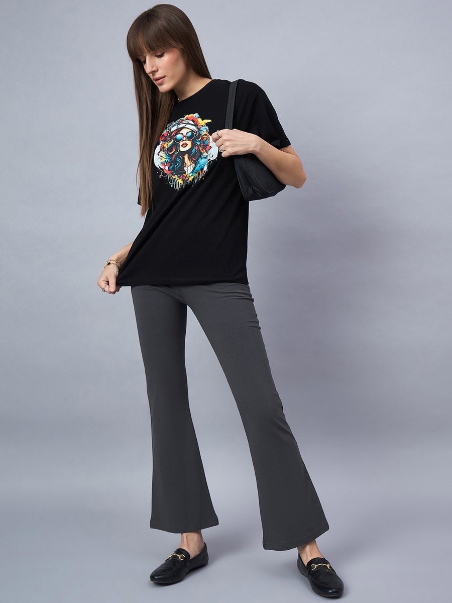 Woman wears 98degreenorth Black Typography Printed Cotton T-shirt, gray flared pants, and black loafers on gray backdrop.