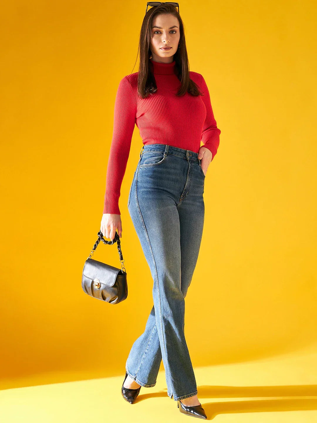 Woman in a 98degreenorth bright red ribbed turtleneck sweater pairs it with blue jeans and heels, holding a black purse.