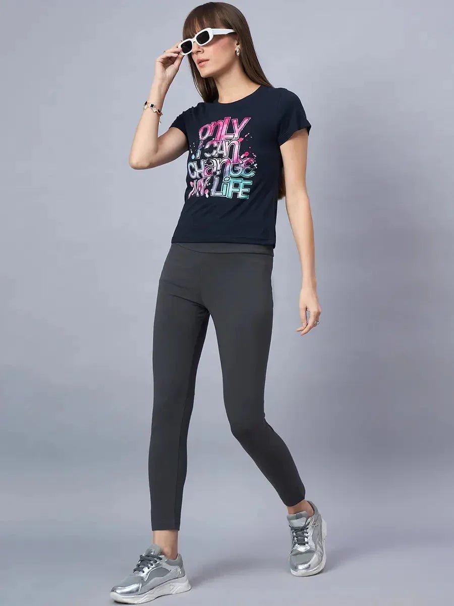 Woman in a 98degreenorth soft cotton typography t-shirt, gray leggings, silver sneakers, and white sunglasses poses.