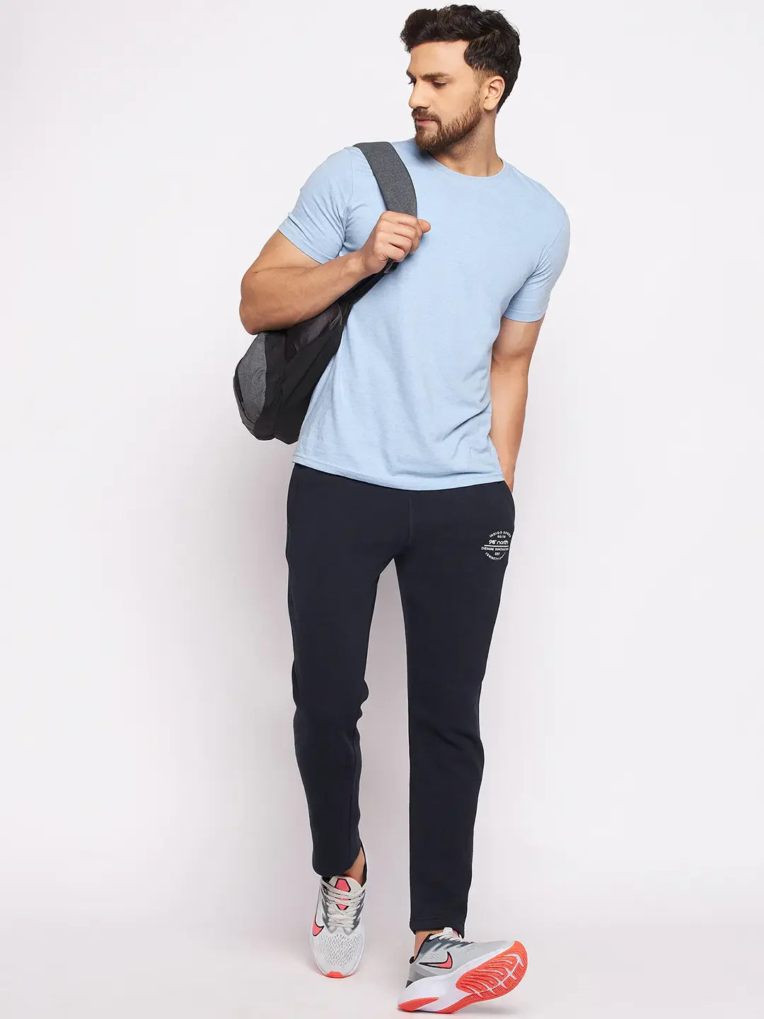 Man wears 98degreenorth Men’s Navy Fleece Trackpants, light blue tee, sneakers, and a backpack against a white background.