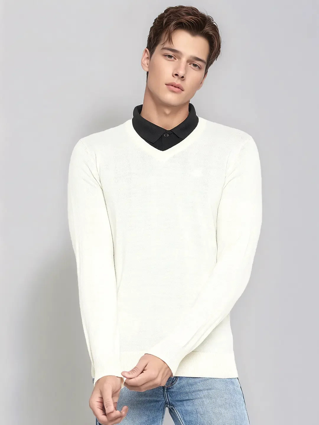 A man wears the 98degreenorth Men’s Off-White Merino Wool V-Neck Sweater with blue jeans against a gray backdrop.