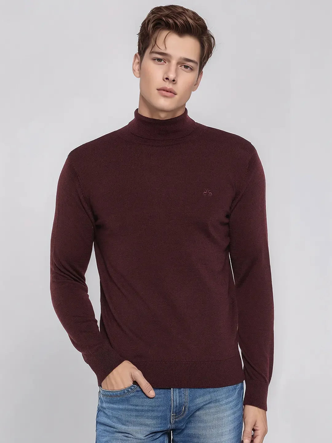 A man in a 98degreenorth men’s wine merino wool turtle neck sweater and blue jeans stands against a gray background.