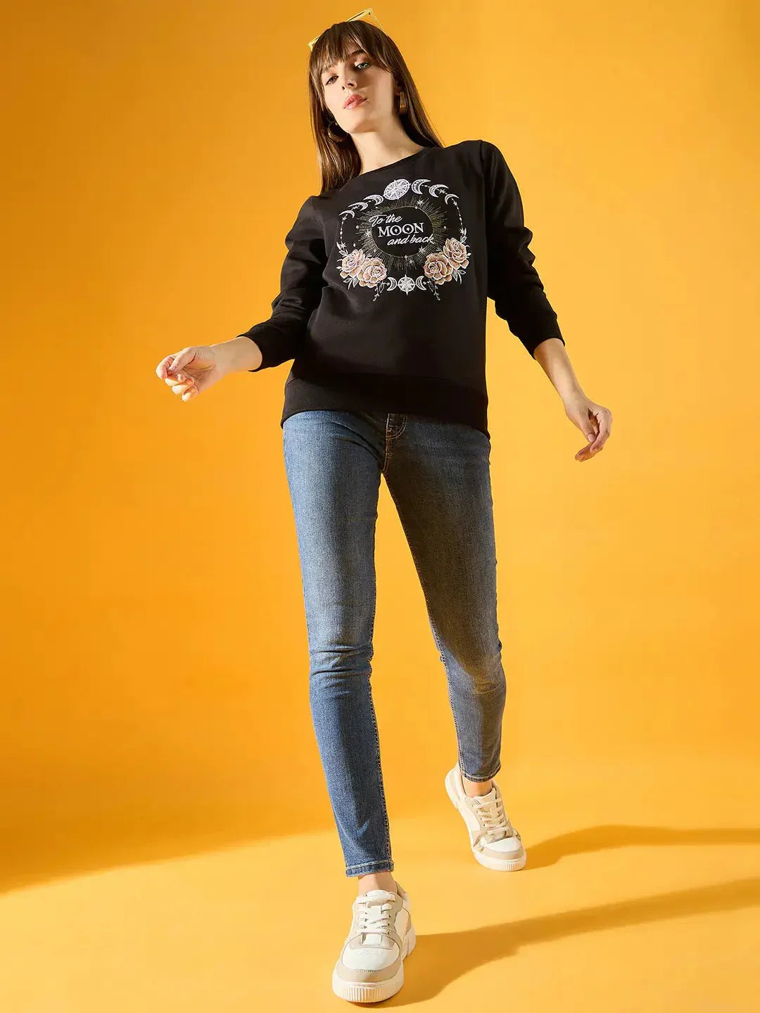 A woman poses against a yellow background, wearing a 98degreenorth Women Graphic Embroidered Black Round Neck Fleece Sweatshirt, blue skinny jeans, and white sneakers.