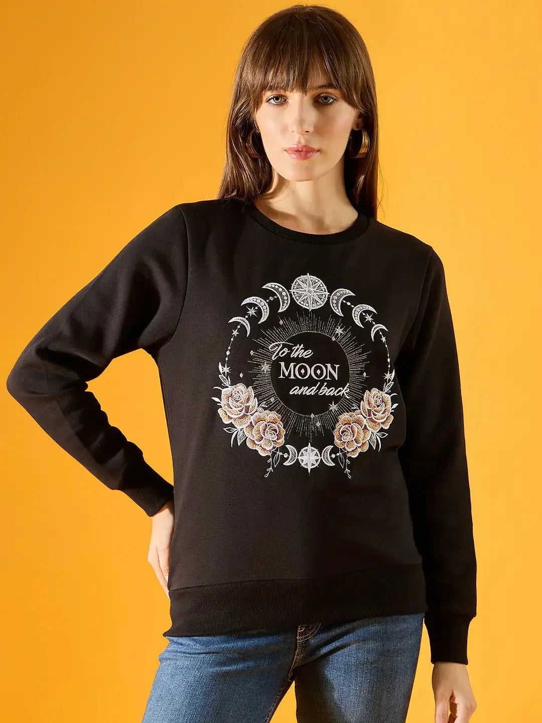 A woman wears the 98degreenorth Women Graphic Embroidered Black Round Neck Fleece Sweatshirt, featuring celestial and floral designs with “To the moon and back” on the front, against a yellow background.