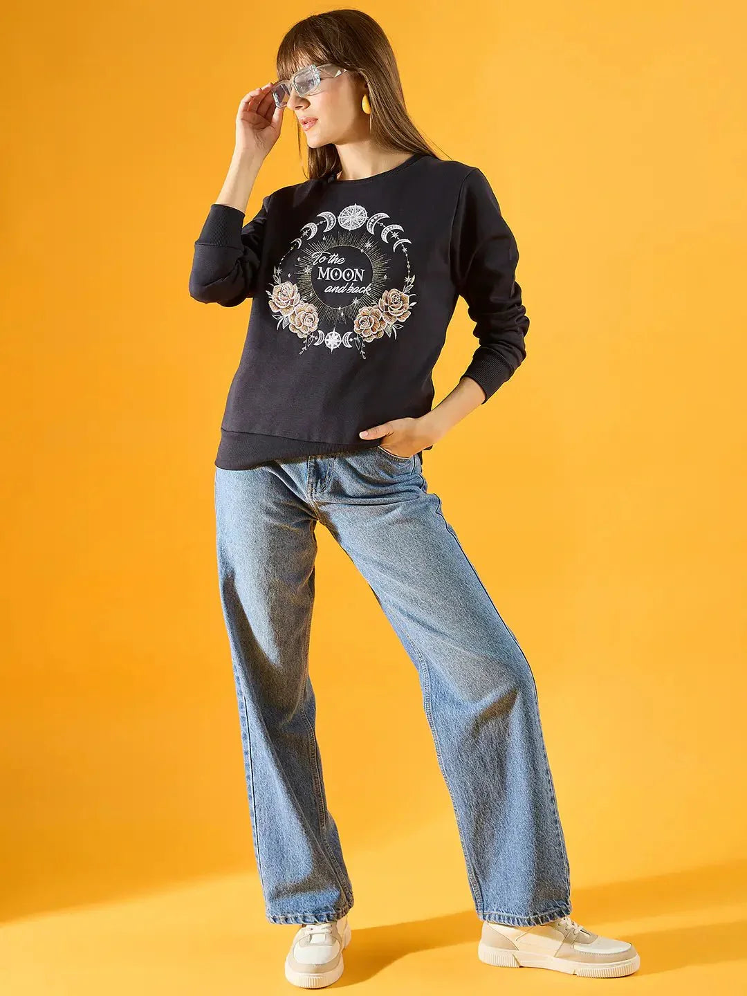 A woman poses against a yellow background in a 98degreenorth Women Graphic Embroidered Navy Round Neck Fleece Sweatshirt, light blue wide-leg jeans, white sneakers, and sunglasses. One hand is in her pocket while the other adjusts her sunglasses.