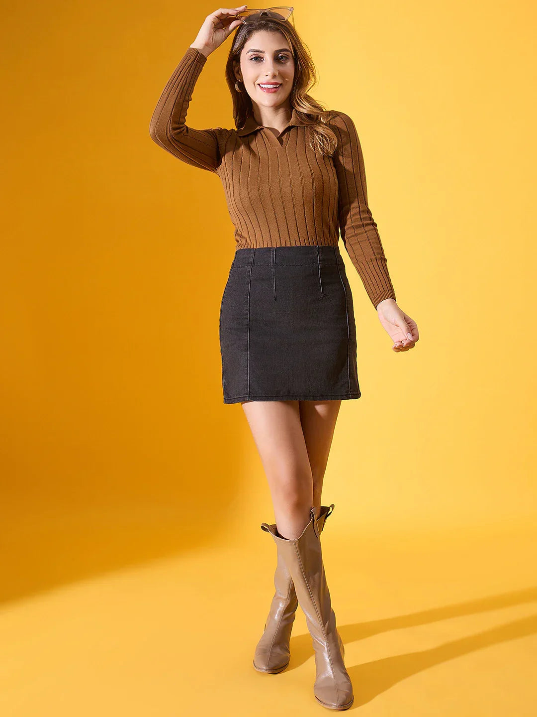 Woman in a 98degreenorth brown ribbed collar neck sweater and black mini skirt poses against a yellow background.