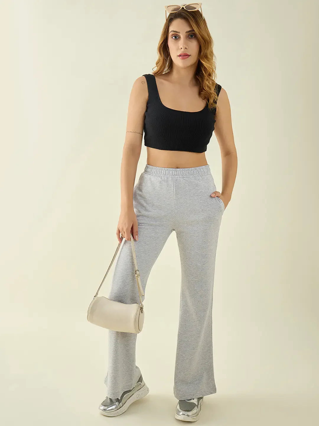 Woman in black crop top and 98degreenorth Light Grey Flared Fit Bootcut Trousers stands with silver sneakers and beige bag.