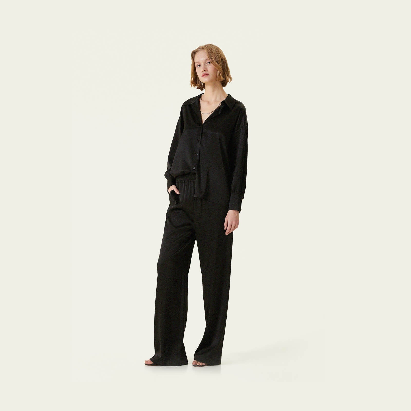 A person stands against a plain light background, wearing a loose-fitting black blouse and wide-leg black trousers with open-toe sandals.