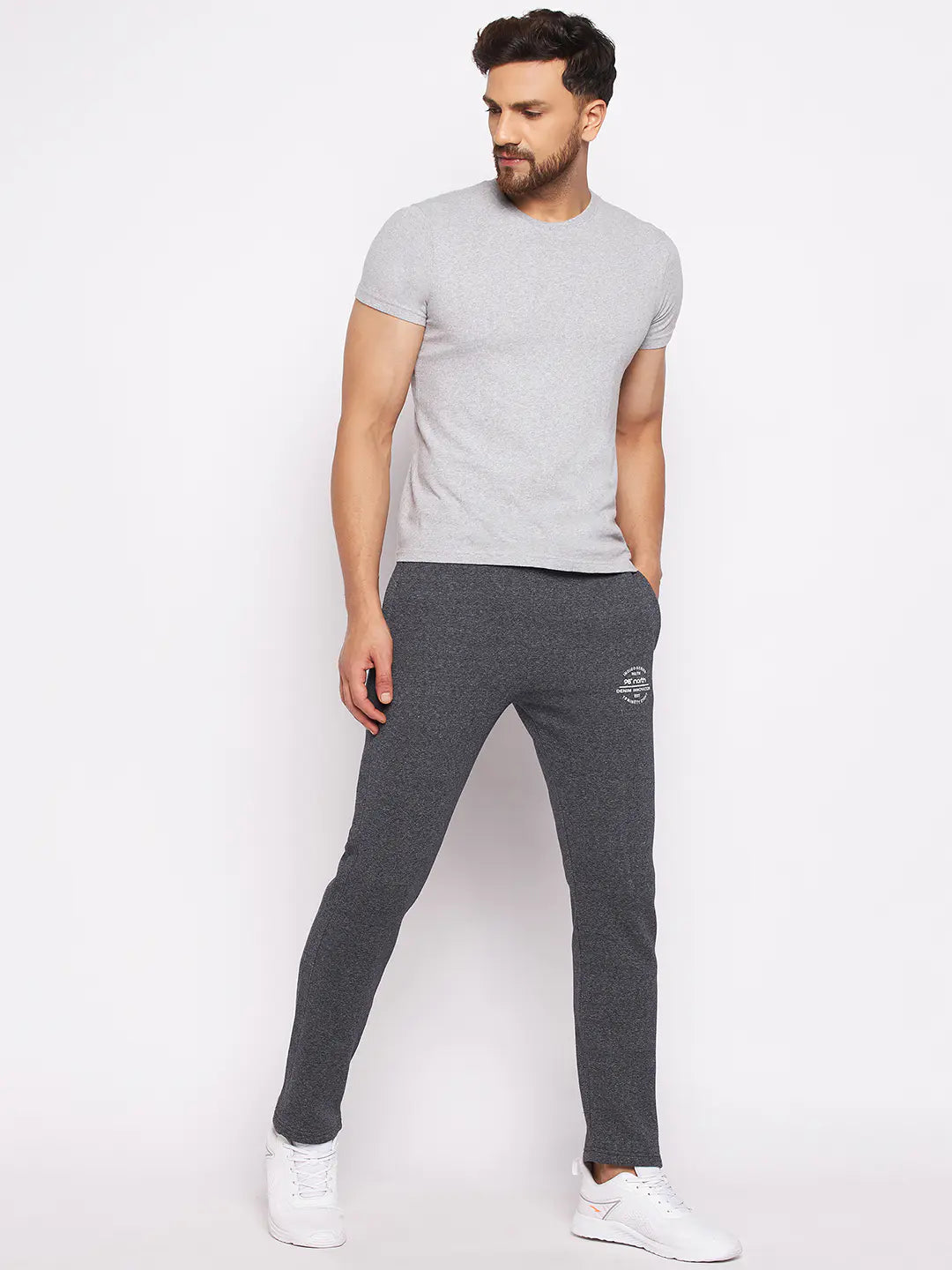 Man wearing a fitted light grey t-shirt, dark grey joggers, and white sneakers, standing against a plain white background with one hand in his pocket.