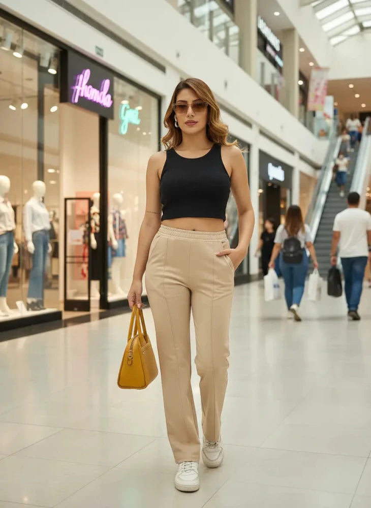 A woman wearing a black crop top, beige pants, and white sneakers walks confidently in a shopping mall, holding a yellow handbag. Mannequins and shoppers are visible in the background.