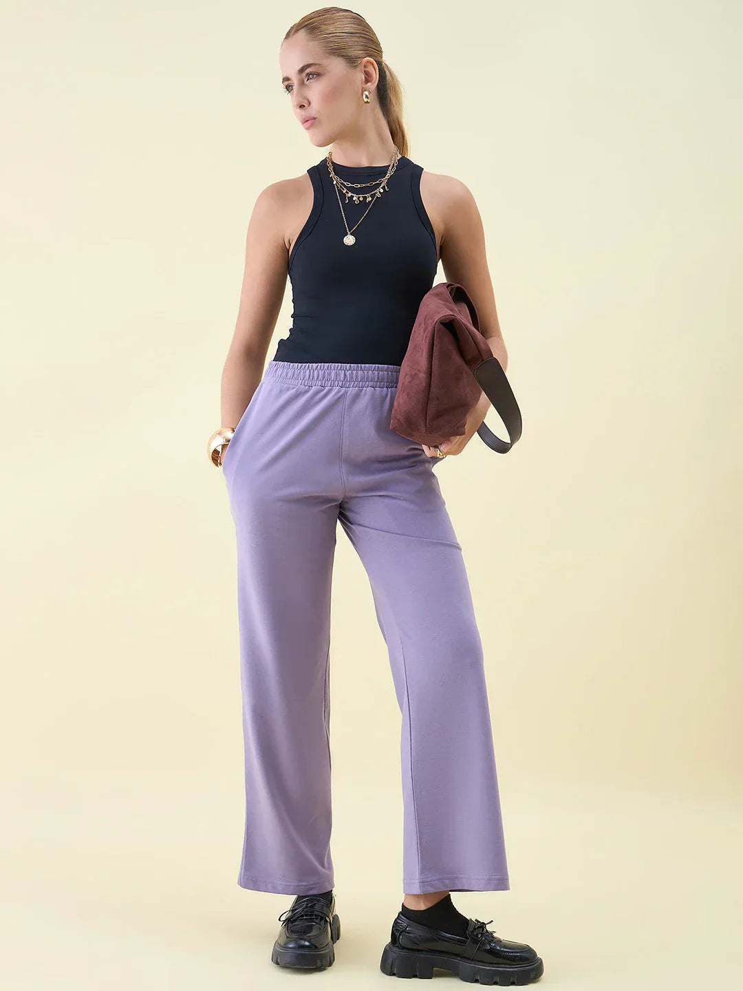 A woman wearing a black sleeveless top, lilac wide-leg pants, and black shoes stands holding a brown bag. She accessorizes with layered necklaces and earrings against a light beige background.