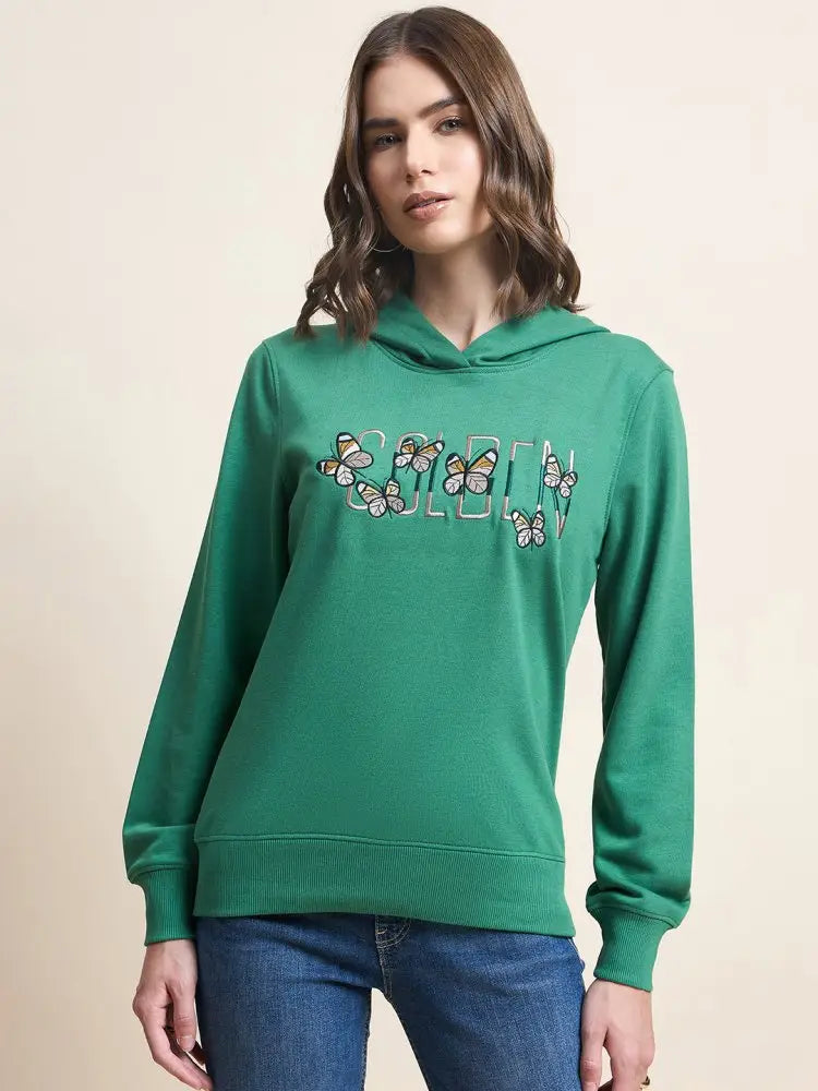A woman wearing a green hoodie with butterfly designs and the word GOLDEN on the front, paired with blue jeans, standing against a plain beige background.