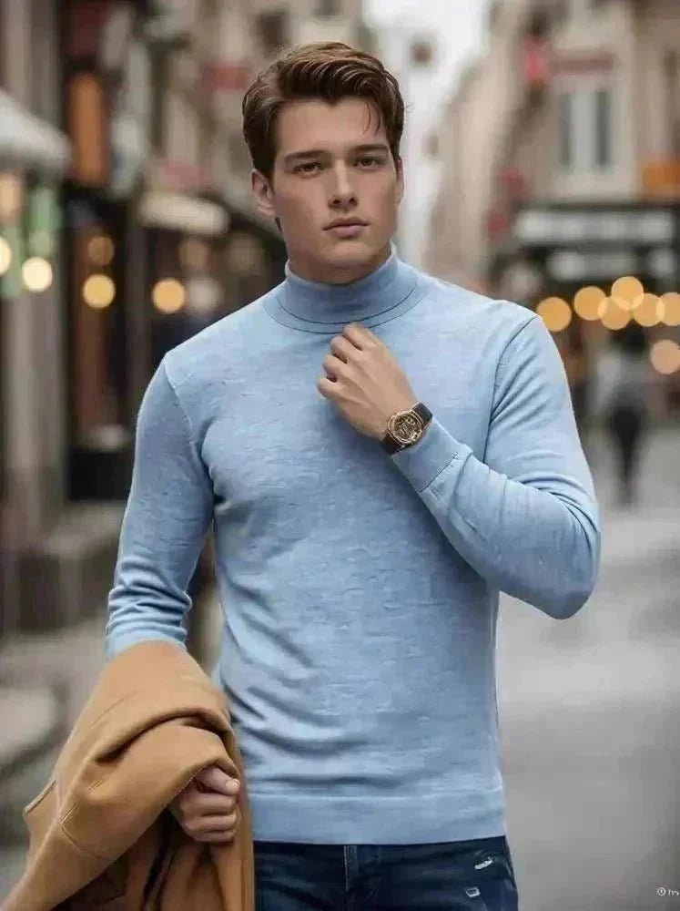 A man walks on a cobblestone street in winter, wearing a burgundy turtleneck, blue jeans, and brown boots, holding a tan coat. Snow is on the ground, and outdoor lamps and buildings line the street.