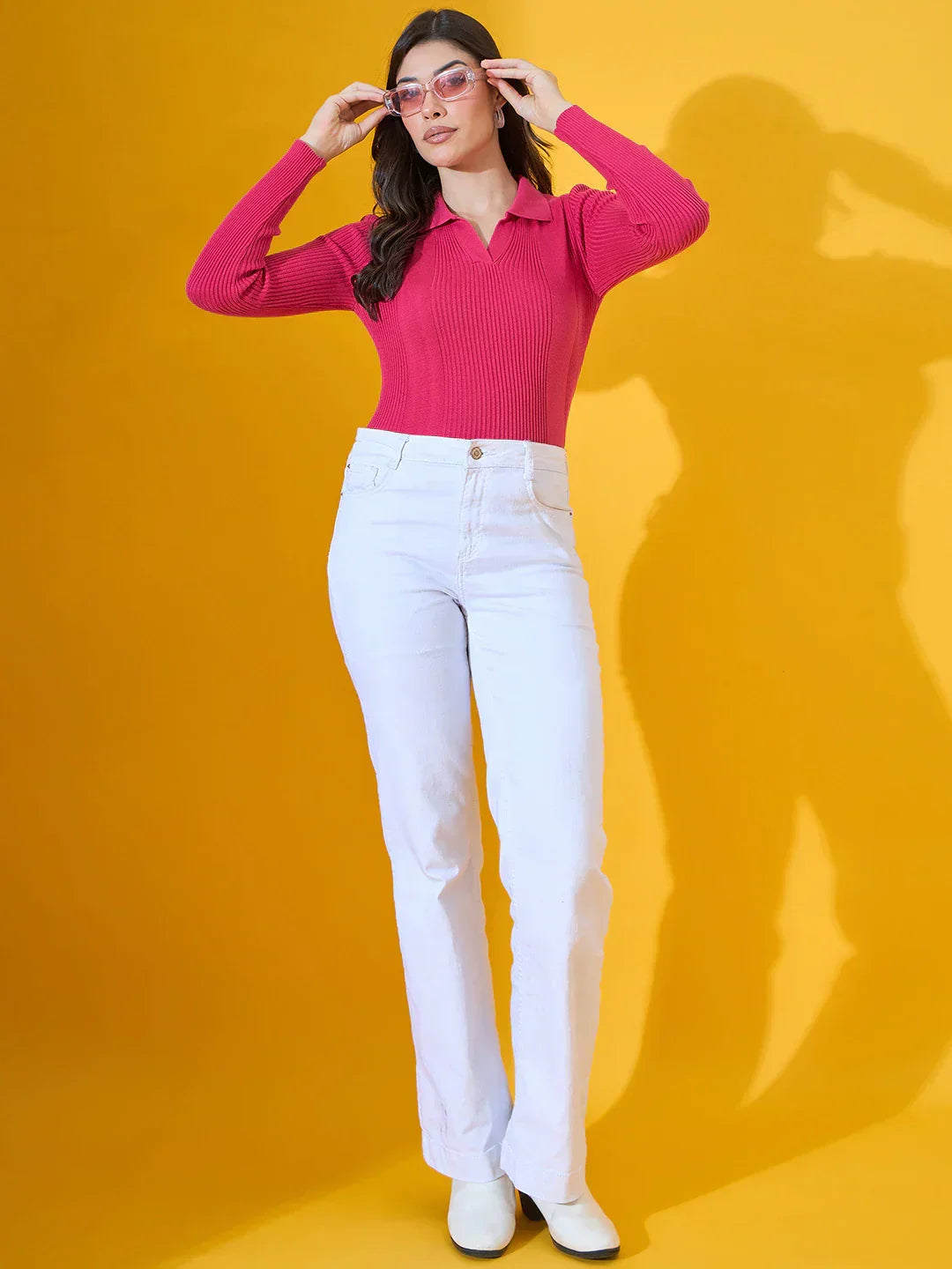 A woman stands against a yellow background, wearing a bright pink long-sleeve top, white high-waisted pants, white shoes, and sunglasses while holding her glasses with both hands.