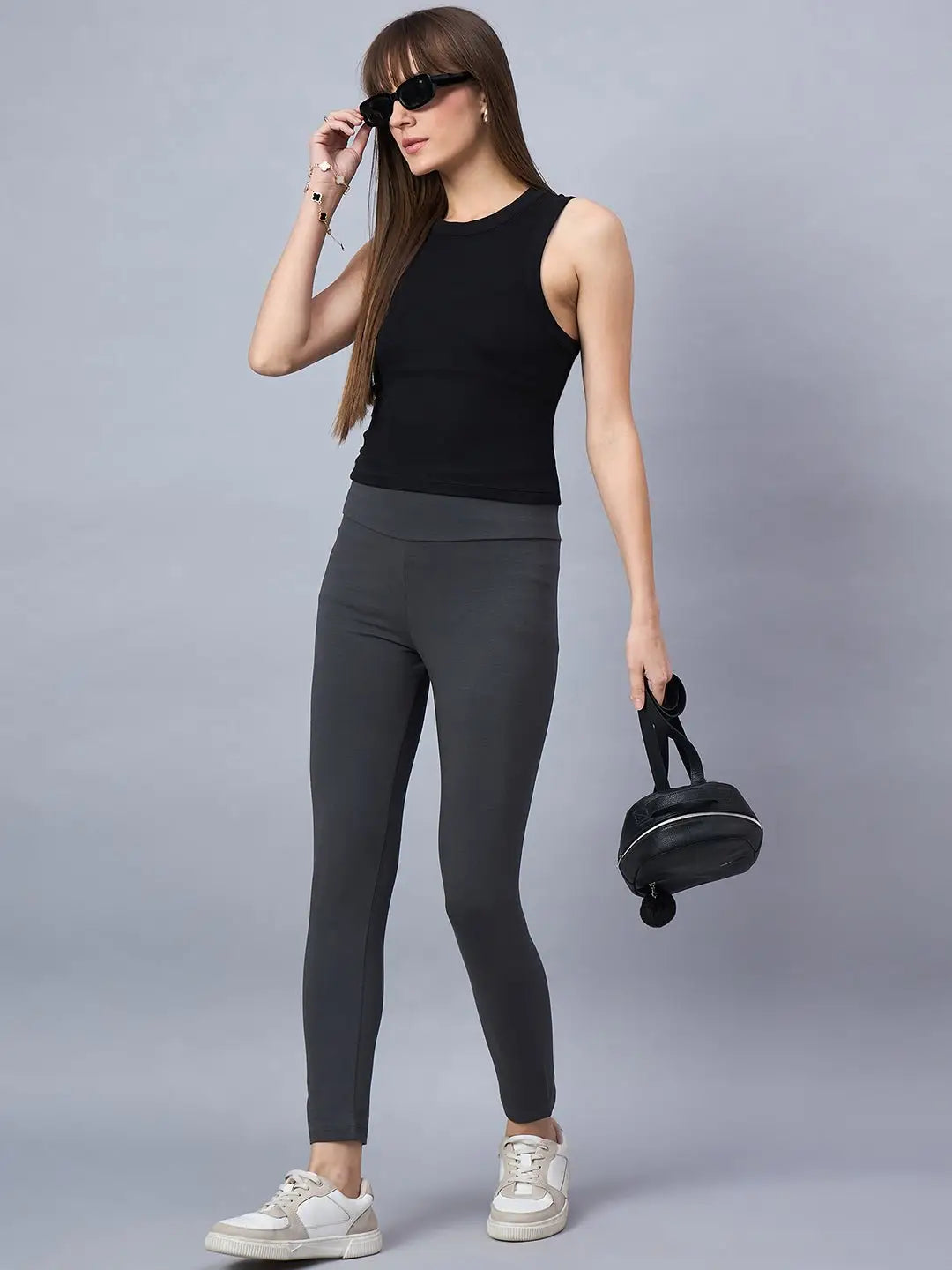 Woman wearing a black sleeveless top, dark gray leggings, white sneakers, and sunglasses, holding a small black round bag, standing against a plain gray background.