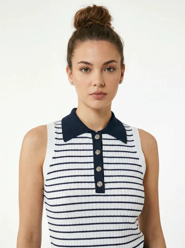 A woman with brown hair in a bun wears a sleeveless white top with navy blue horizontal stripes, a navy blue collar, and a button placket. She stands against a plain light background.