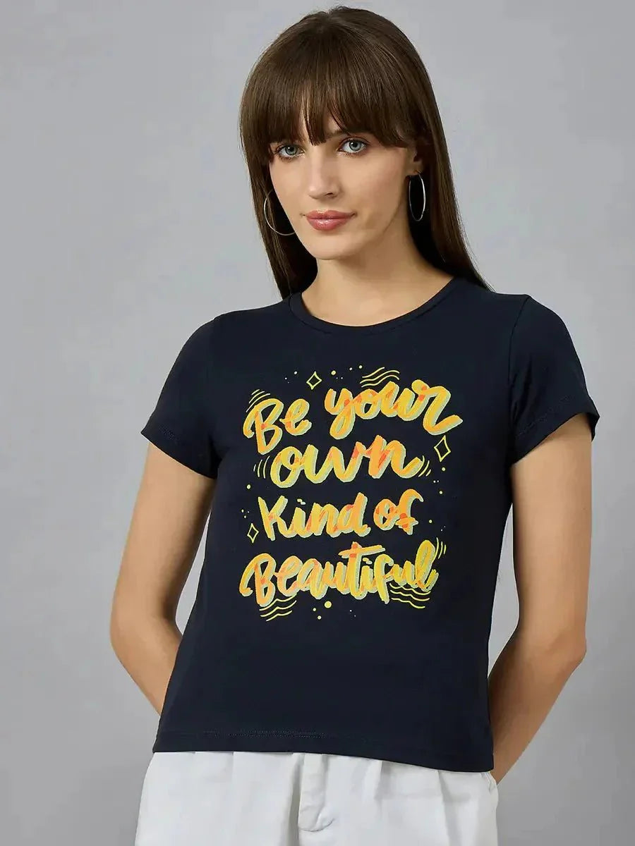A woman with straight brown hair and bangs wears a black t-shirt with the yellow text Be your own kind of beautiful. She stands against a plain gray background, looking at the camera.