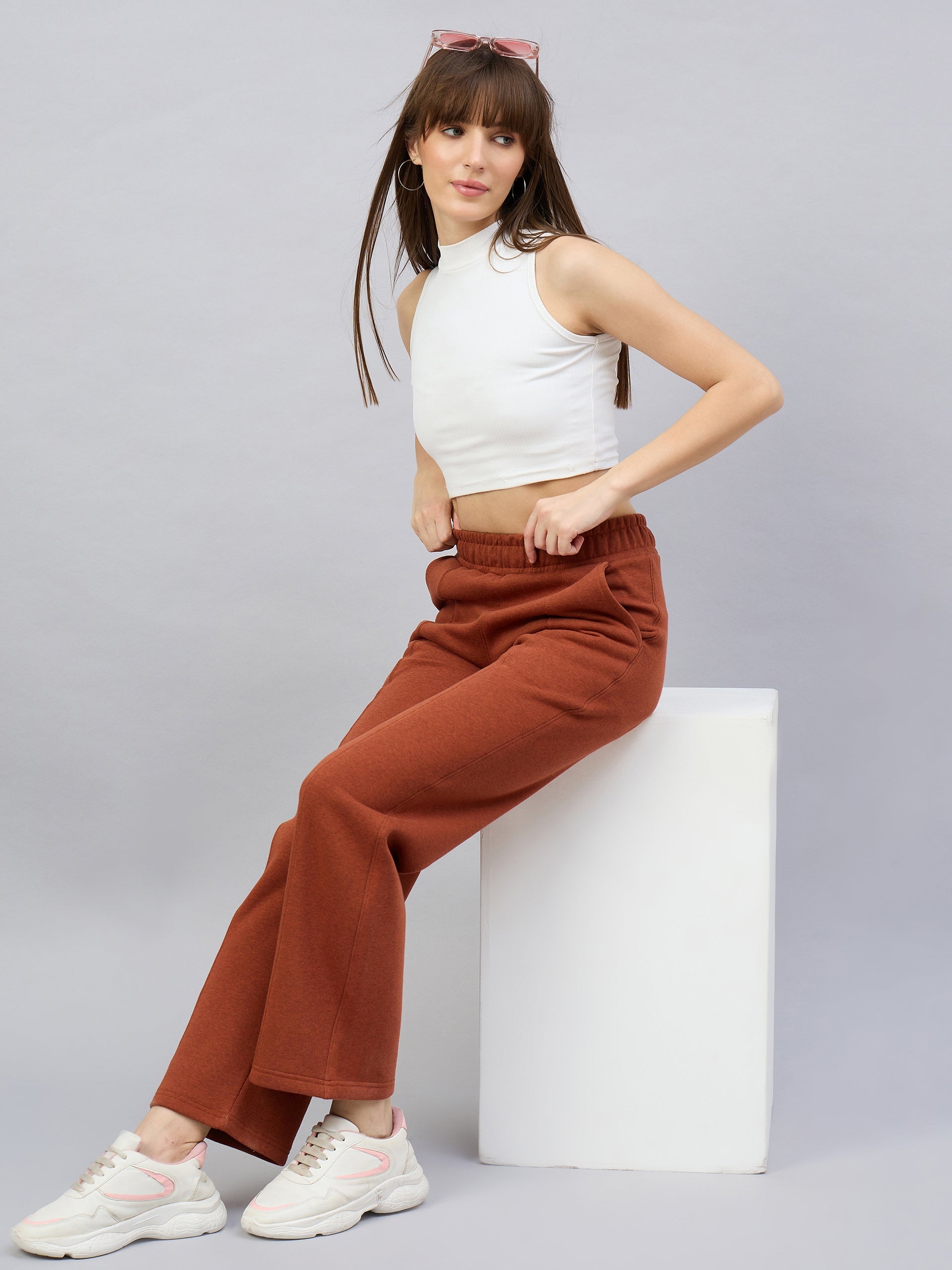 Woman in a white sleeveless crop top, rust-colored pants, and sneakers sits on a white block against a gray background.