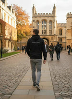 A man in a 98degreenorth Black Fleece Zipper Hoodie walks down a cobblestone path at a historic university campus.