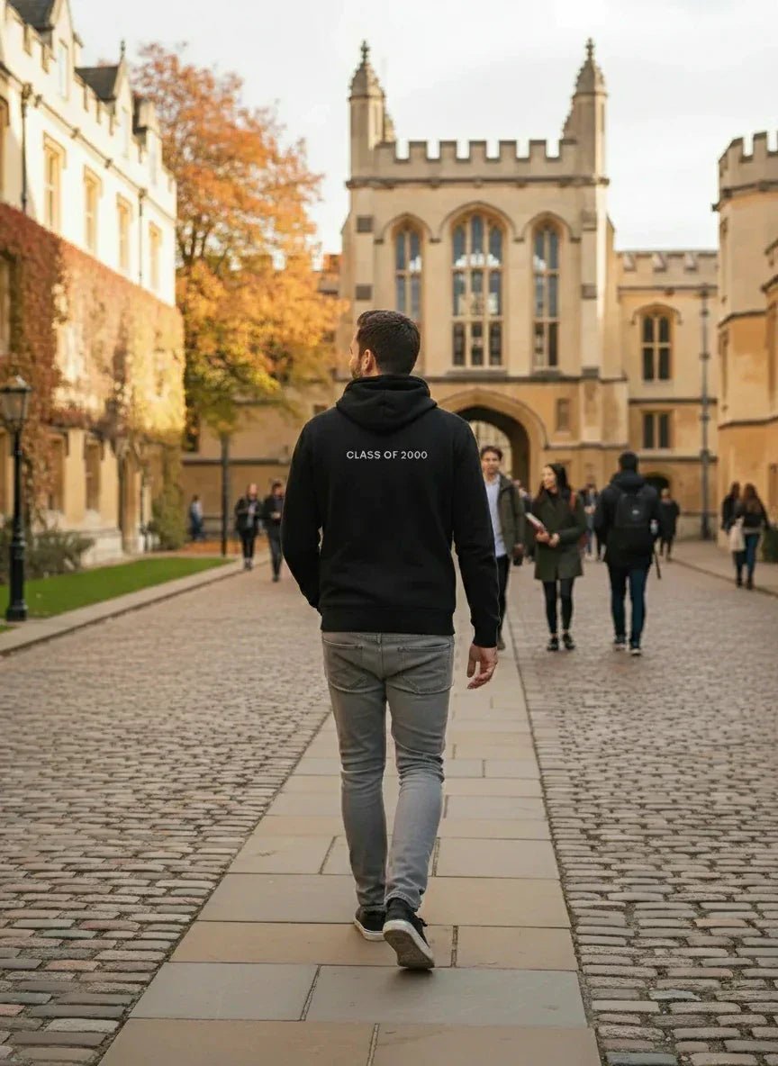 A man in a 98degreenorth Black Fleece Zipper Hoodie walks down a cobblestone path at a historic university campus.