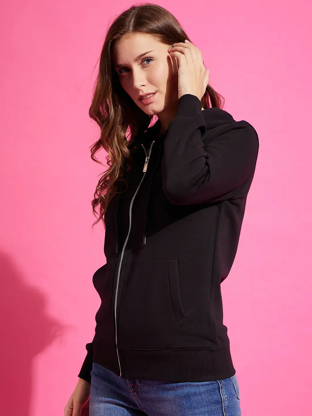 Woman with long brown hair in a 98degreenorth black fleece zipper hoodie sweatshirt, posing against a pink background.
