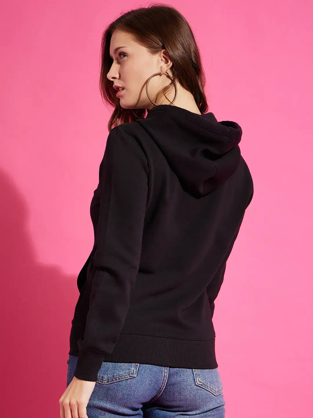 Woman wears 98degreenorth black fleece zipper hoodie sweatshirt and blue jeans, facing away against a pink background.