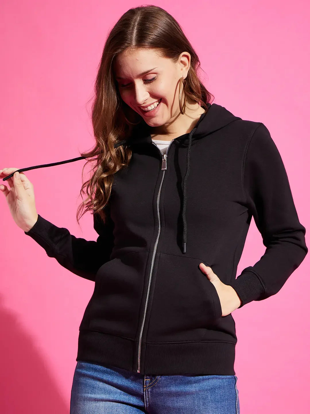 Woman in a 98degreenorth Black Fleece Zipper Hoodie smiles, holding the string, against a pink background.