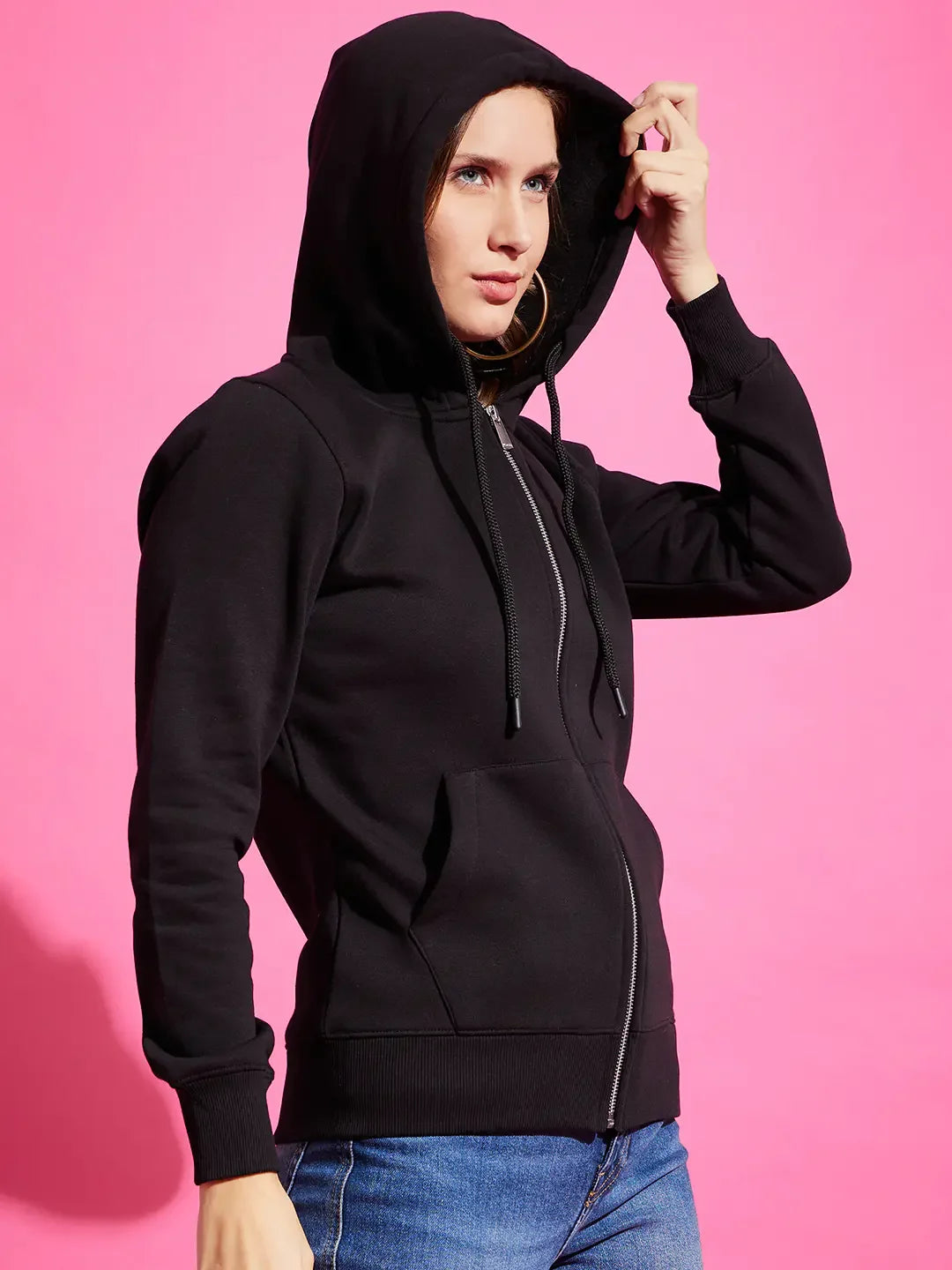 Woman wears 98degreenorth black fleece zipper hoodie with blue jeans, standing against a bright pink background.