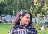 Woman in a black patterned outfit standing outdoors, smiling, with trees and people sitting on grass in the background.