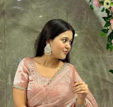 Woman in a pink saree with large earrings, looking to the side, standing near pink flowers against a marble wall.