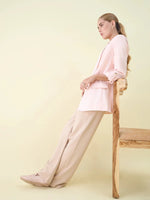 Woman in beige wide leg pintuck trackpants by 98degreenorth lounges on a chair against a pale yellow background.