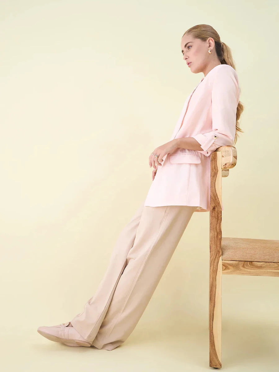 Woman in beige wide leg pintuck trackpants by 98degreenorth lounges on a chair against a pale yellow background.