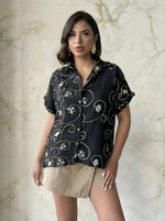 Woman in a 98degreenorth Black Floral Embroidered Linen Shirt and beige skirt stands by a light marble wall.