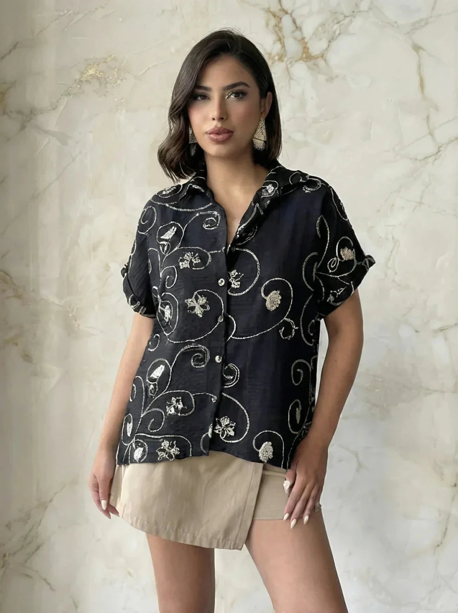Woman in a 98degreenorth Black Floral Embroidered Linen Shirt and beige skirt stands by a light marble wall.