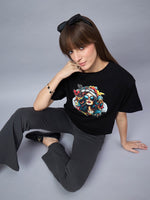 Woman wears a 98degreenorth Black Typography Printed Cotton T-shirt, gray pants, and black shoes, looking up at camera.