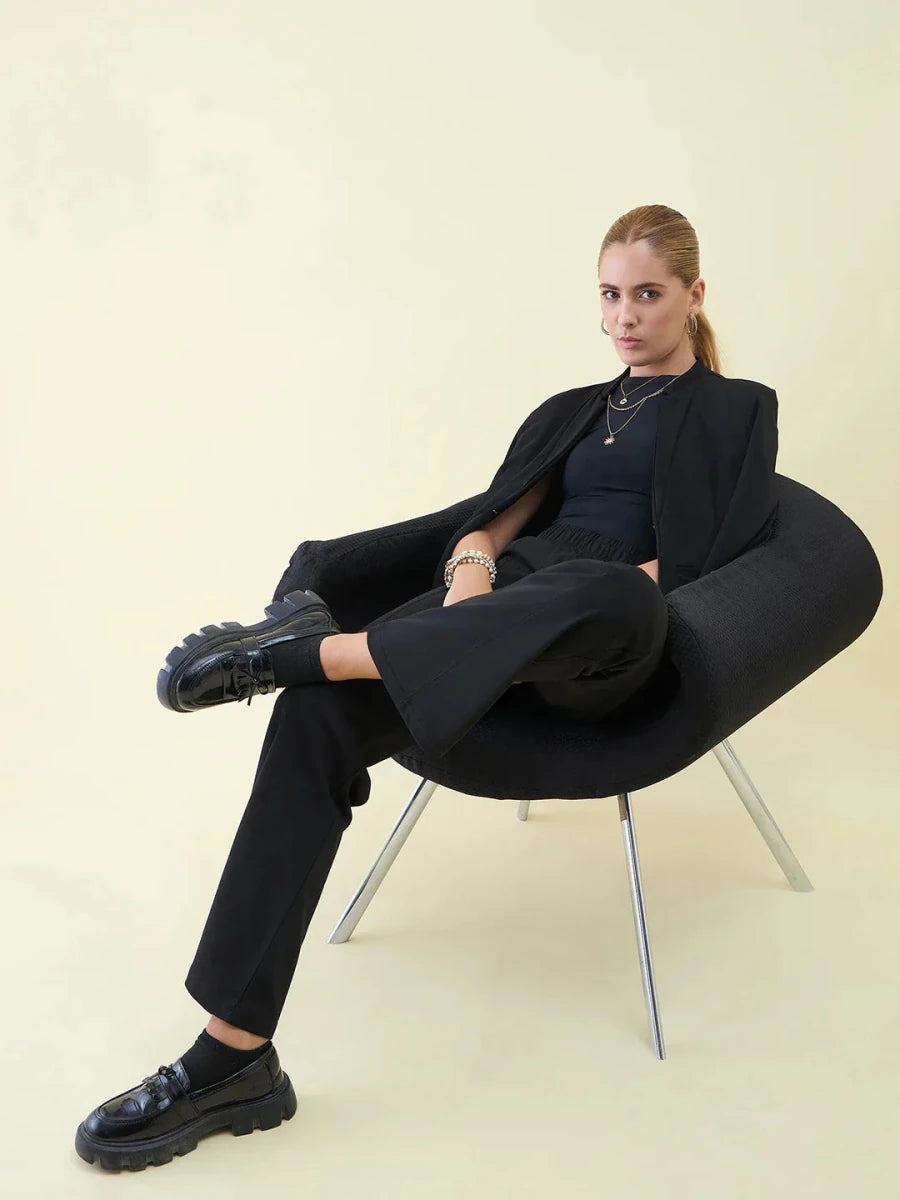 A woman in 98degreenorth Black Wide Leg Pintuck Trackpants sits confidently in a modern black chair.