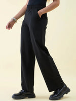 Model in 98degreenorth Black Wide Leg Pintuck Trackpants and chunky shoes, styled with bracelets on a beige background.