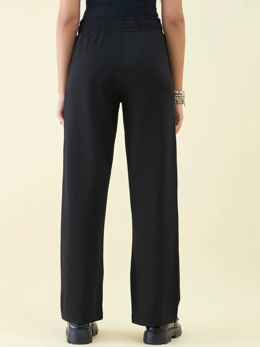 Model wears 98degreenorth Black Wide Leg Pintuck Trackpants from Luxe Lounge, seen from the back on a beige background.