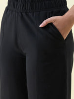 Model wears 98degreenorth Black Wide Leg Pintuck Trackpants, hand in pocket, shown waist to mid-thigh.