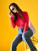 Woman wears 98degreenorth bright red women’s ribbed turtleneck sweater against a yellow background, adjusting sunglasses.