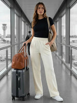Woman in a black top and 98degreenorth Butter Wide Leg Pintuck Trackpants stands at the airport with suitcase and brown bag.