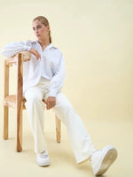 Woman in Butter Wide Leg Pintuck Trackpants by 98degreenorth sits on a wooden chair, looking to the side.