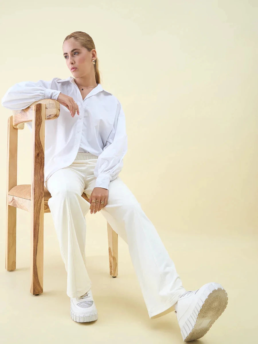 Woman in Butter Wide Leg Pintuck Trackpants by 98degreenorth sits on a wooden chair, looking to the side.