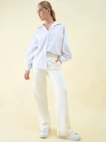Person in a white oversized shirt and Butter Wide Leg Pintuck Trackpants by 98degreenorth stands against a pale yellow background.