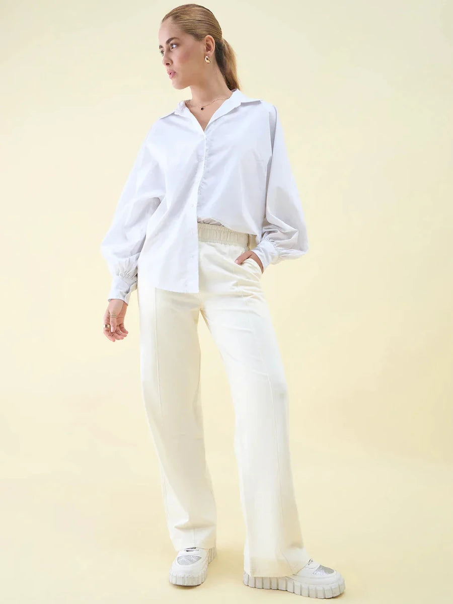 Person in a white oversized shirt and Butter Wide Leg Pintuck Trackpants by 98degreenorth stands against a pale yellow background.