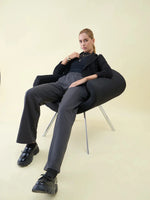 Woman in 98degreenorth Charcoal Wide Leg Pintuck Trackpants sits confidently in a black chair against a light background.