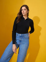 Woman in a 98degreenorth Classic Black Ribbed Polo-Neck Knit Top and blue jeans poses against a yellow background.