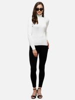 Woman in 98degreenorth Classic White Ribbed Turtle Neck Sweater, black leggings, sunglasses & sandals vs. plain backdrop.