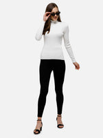 Woman wears 98degreenorth Classic White Ribbed Turtle Neck Sweater, black sunglasses, leggings, and heels, smiling.