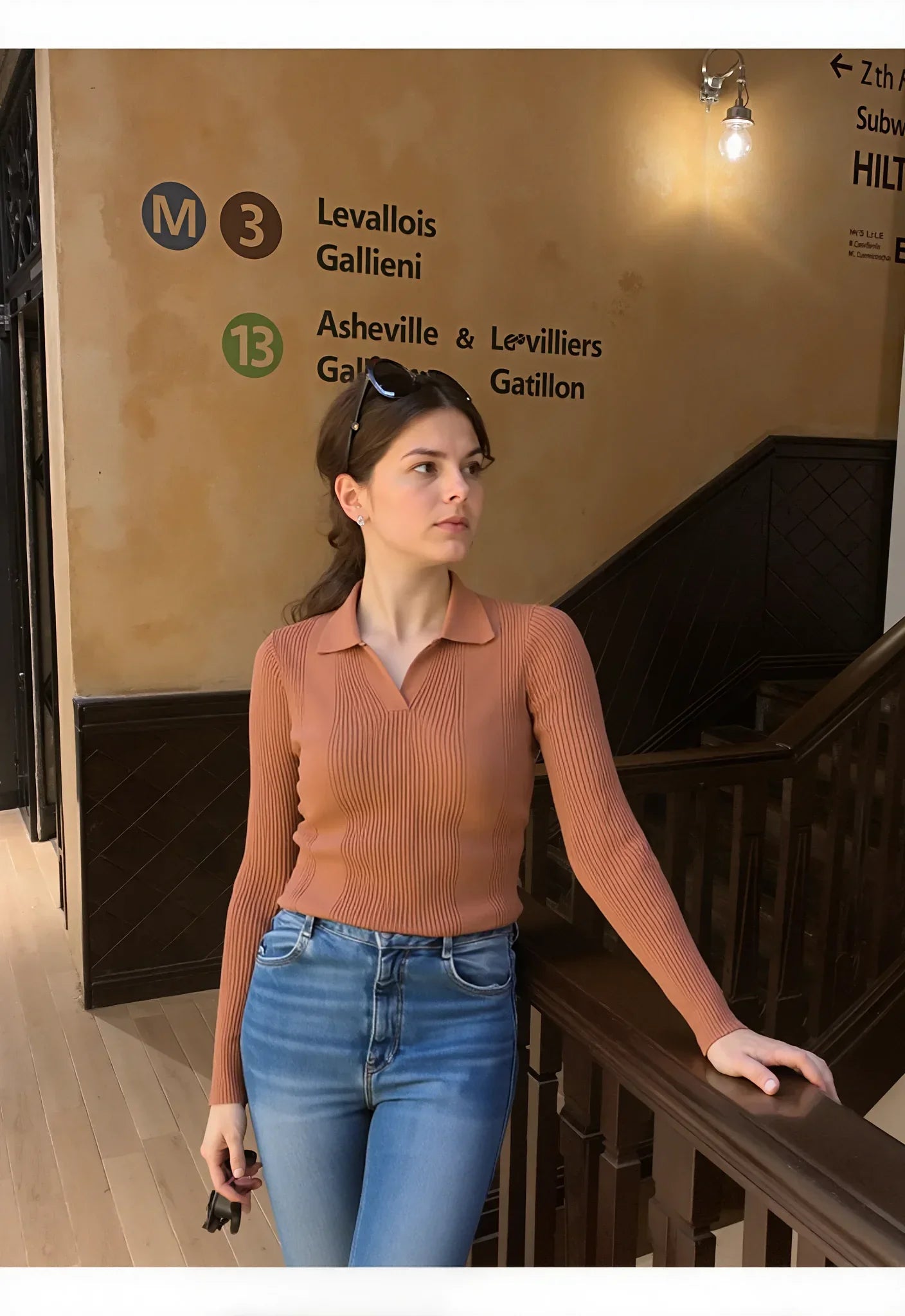 Woman wears 98degreenorth Dark Camel Ribbed Knit Collared Sweater, standing near stairs with subway signs behind her.